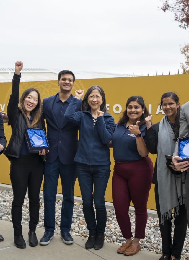 Group of individuals standing in front of the UC Davis sign with their hands raised celebrating